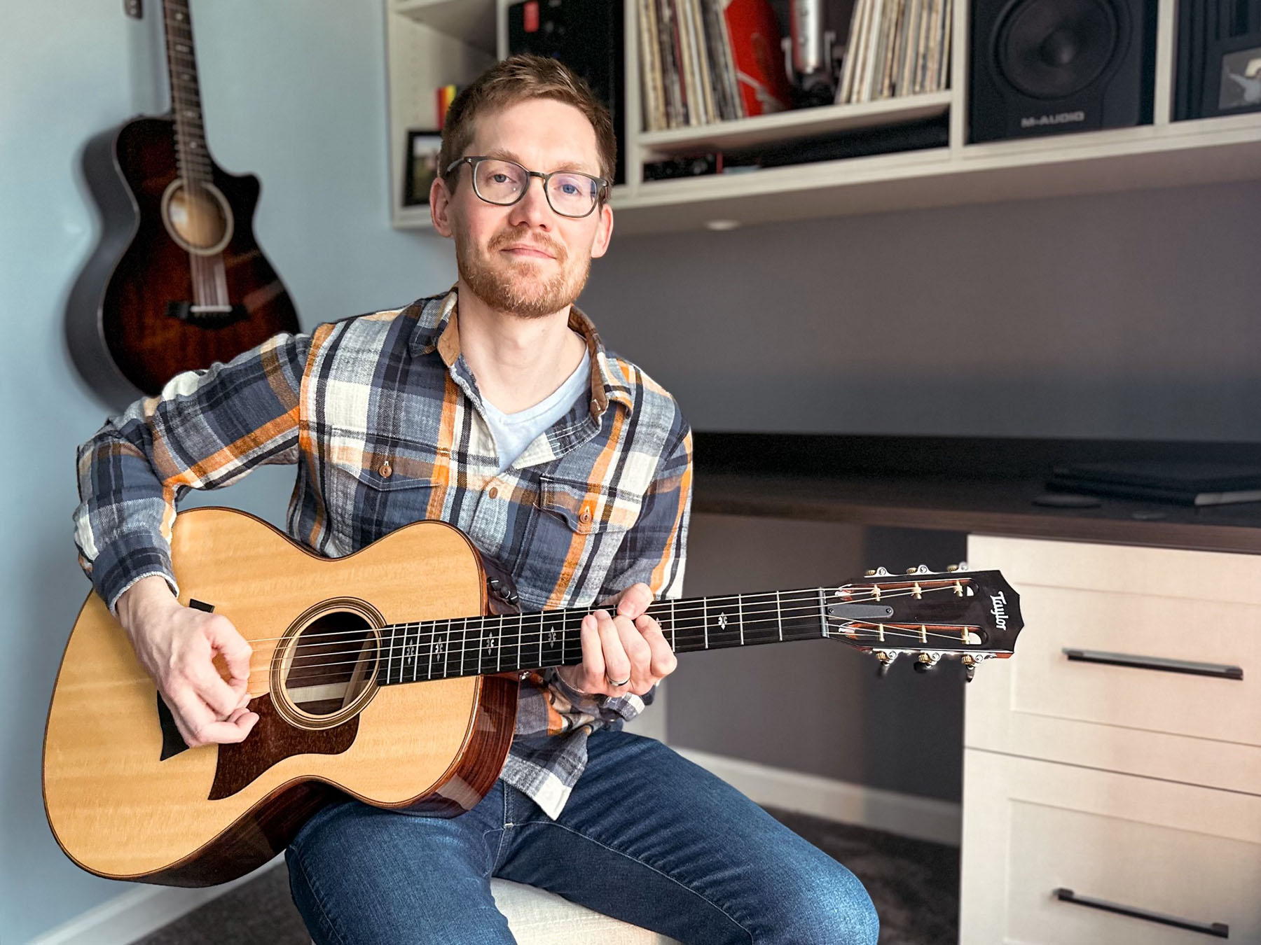 A man in jeans and a flannel shirt sits with an acoustic guitar