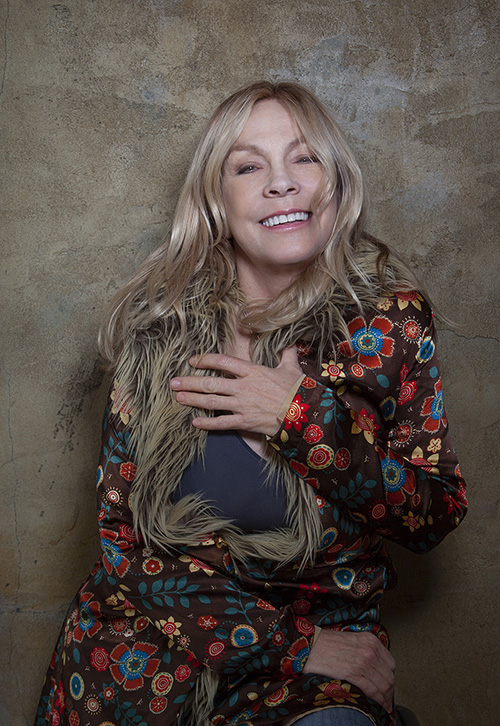 Sporting long blonde hair and a colorful floral jacket, singer Rickie Lee Jones leans back on a wall and smiles. Photo by Astor Martin.